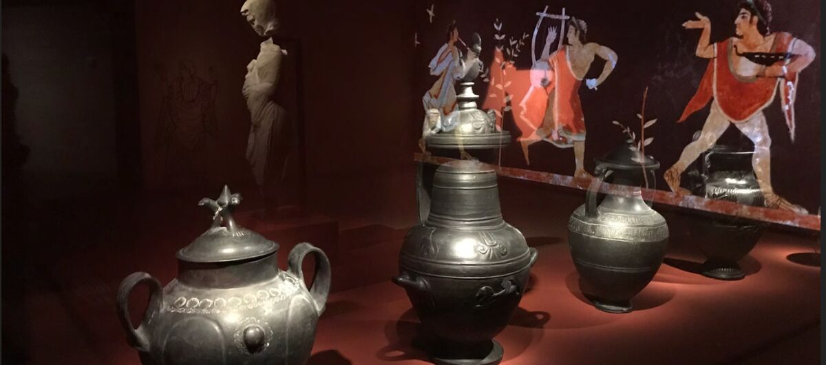 Foto von drei metallischen Gefäßen unterschiedlicher Form. Sie stehen auf einer dunkelroten Oberfläche. Der Hintergrund ist sehr dunkel. Angeleuchtet stehen dort eine Statur sowie ein Bild, das eine Szene mit drei Menschen vergangener Zeiten darstellt.