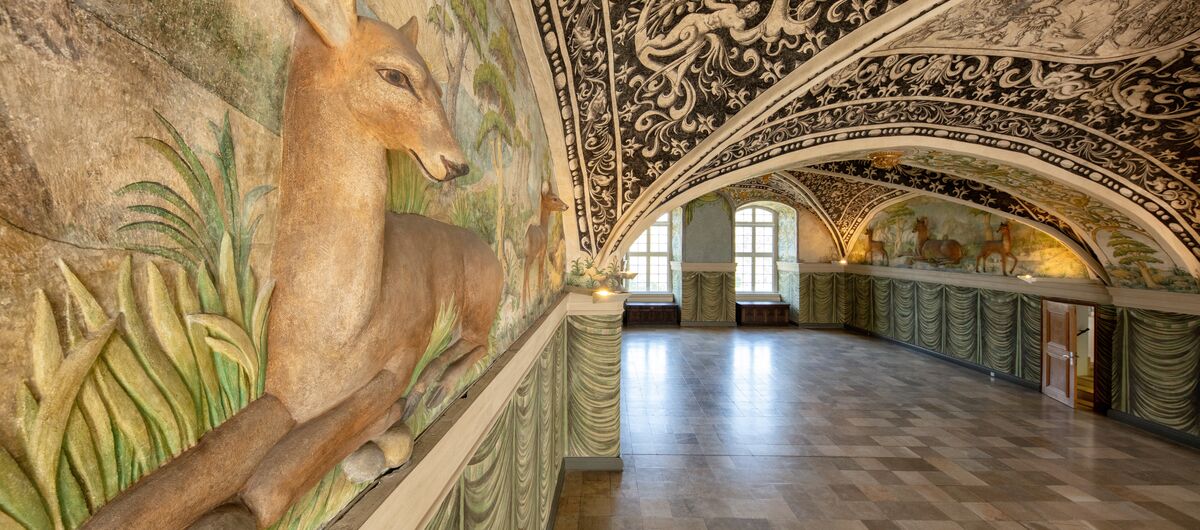 Foto im Hirschsaal des Schloss Gottorf. Die linke Fotoseite zeigt eine dreidimensionale Holzvertäfelung, die zwei Hirsche in der Natur zeigt. Das Holz ist mit sanften Farben bemalt. Der rechte Teil des Fotos zeigt das kunstvoll bemalte Gewölbe des Saals.