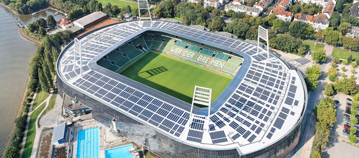 Luftaufnahme des Weserstadions. Das Stadion ist oval, die Besucherplätze überdacht. Auf dem Stadiondach stehen vier Fluchtlichtinstallationen. Links unter dem Stadion ist ein Freibad mit zwei Schwimmbecken zu erkennen. Links nks daneben fließt die Weser.