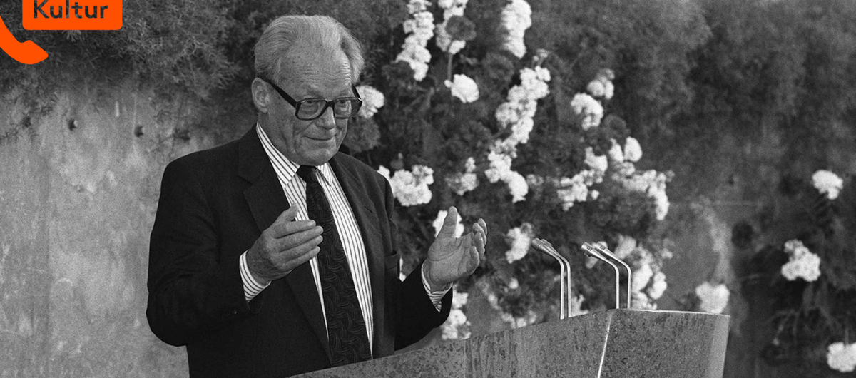 Schwarz-weiß Foto von Willy Brandt im Anzug während einer Rede. Er steht an einem Pult mit schwarzer Brille, Blick in Richtung Zuhörende und Händen auf Brusthöhe. Hinter ihm eine Steinwand an der Hecke und Blumen hinabranken.