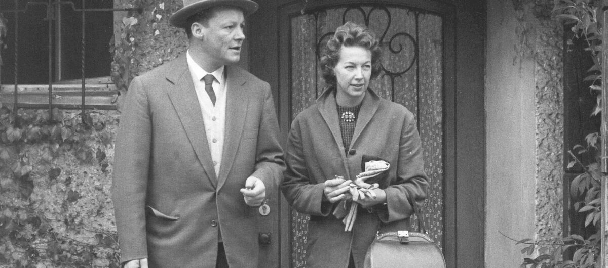 Schwarz-weiß Foto von Willy und Rut Brandt Ende der 50er Jahre. Sie stehen vor einer Haustür und schauen von der betrachtenden Person aus neutral nach rechts. Weinblätter und Efeu hängen über ihnen vom Haus hinunter.