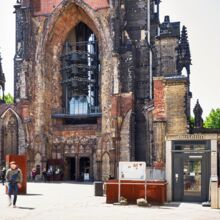 Frontaler Blick auf die Überreste von St. Nikolai mit Glocken im Kirchenschiff in rot-beigen Stein mit schwarzen Verfärbungen. Moderne Bauelemente, wie die Eingangstür zum Museum, fügen sich in den alten Bau ein.