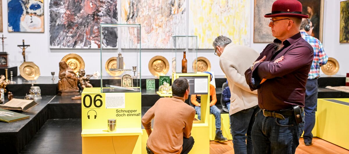 Ausstellungsansicht zu "Hör mal hin!". Besuchende - teils mit Kopfhörern auf dem Ohr - schauen sich Objekte in Vitrinen und auf Podesten sowie Gemälde an Wänden an. Die Objekte sind teils religiöser Herkunft, die Gemälde abstrakt bis realistisch.