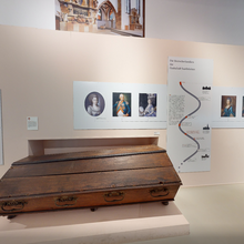 Foto eines Ausstellungsbereichs mit hellen Wänden und Boden. Auf einem flachen Podest steht ein älterer Sarg aus Holz. An der Wand dahinter hängen Informationstafeln mit Texten und älteren Portrait-Gemälden von Frauen und Männern.