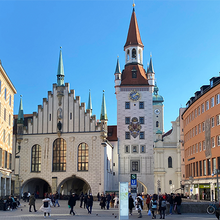 Foto des Marienplatzes in der Münchener Altstadt. Vor blauem Himmel ragen in der Mitte des Bildes ein heller Glockenturm mit einer großen und vier kleinen Turmspitzen sowie ein ebenfalls älteres weißes Gebäude mit vier Turmspitzen in den Himmel.