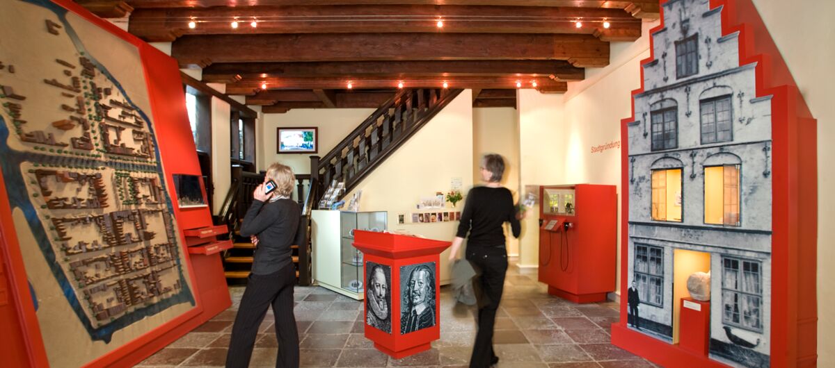 Blick in Ausstellungsraum mit Boden aus Steinplatten. An der Decke sind Fachwerkbalken. Links an der Wand ist ein Stadtmodell, rechts ein großes Fotos eines Hauses zu sehen.  Zwei Personen schauen sich die Ausstellung an.