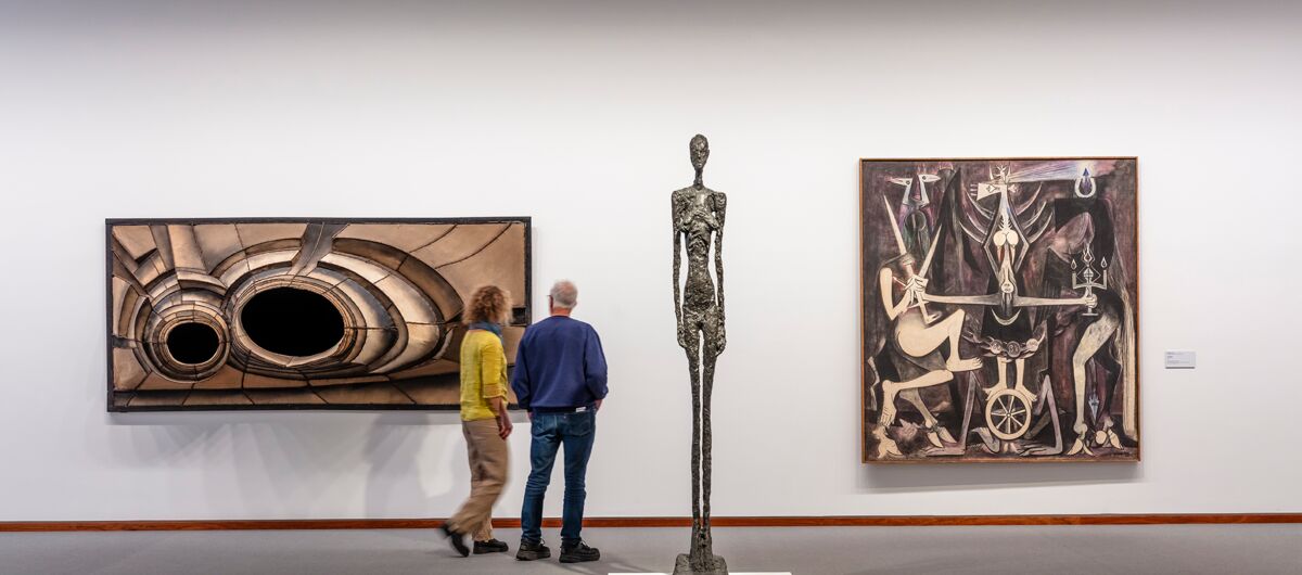 Blick in Ausstellungsraum mit grauem Boden und weißer Wand. Zwei große rechteckige Gemälde hängen an der Wand. Sie sind in Schwarz-, Weiß- und Brauntönen gehalten. Zwischen ihnen steht in den Raum gerückt eine sehr hohe dünne Skulptur.