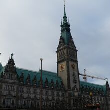 Foto vom Hamburger Rathaus. Auf die Fassade blickend verfügt es mittig über einen hohen verzierten Turm mit Uhr. Die viele große und kleine symmetrisch angeordnete und verzierte Fenster schmücken die Fassade.